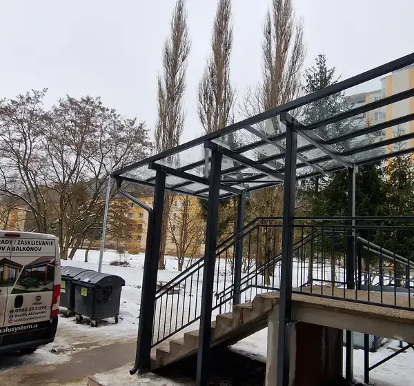 Bočný pohľad na oceľový prístrešok s bezpečnostným sklom pred vchodom do bytovky v Považskej Bystrici, antracitová farba RAL 7016.