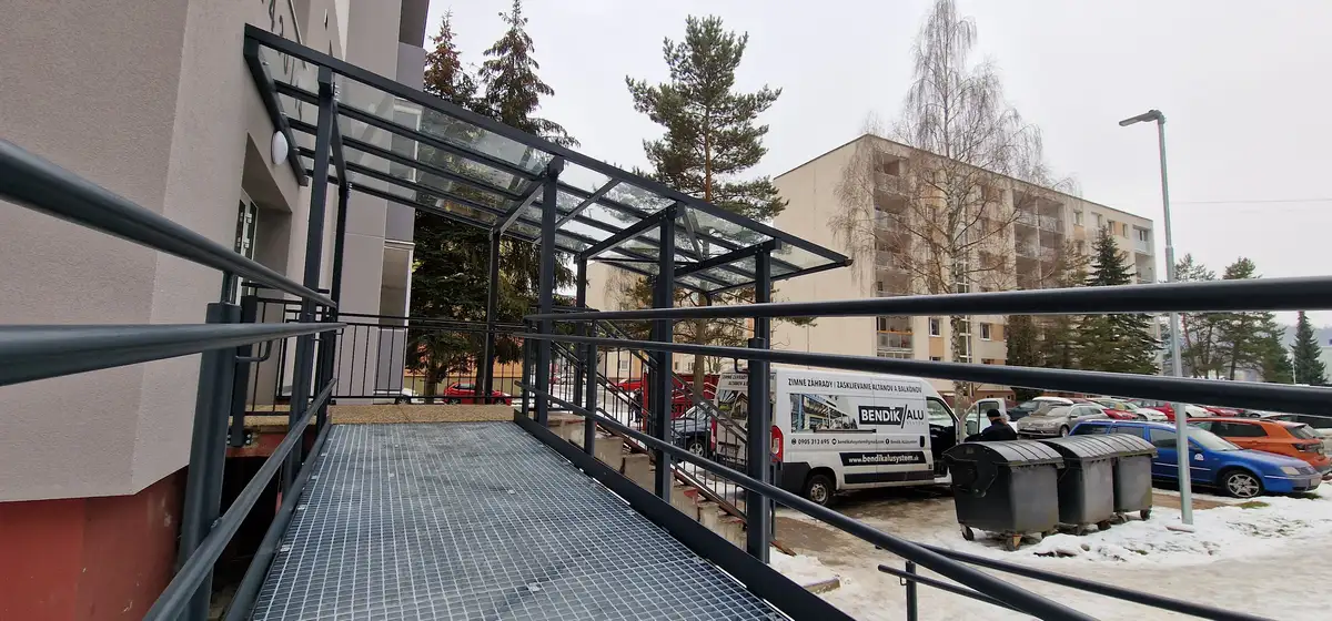 Finálny vzhľad oceľového prístrešku s bezpečnostným sklom a zábradlím pred vchodom do bytovky v Považskej Bystrici, antracitová farba RAL 7016.